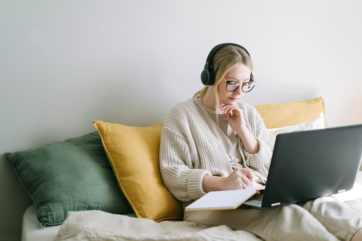 Image of a woman sitting in bed, wearing headphones, using a laptop.